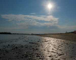 A ground-level view of a wet, pebbly beach foreground leading to a calm sea and a long line of rock breakwaters under a blue sky with white clouds.