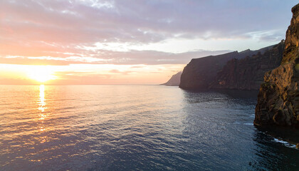 Dramatic sunrise seascape, towering cliffs reflected in calm ocean waters, golden and pink light