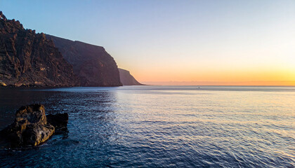 Dramatic sunrise seascape, towering cliffs reflected in calm ocean waters, golden and pink light
