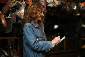 Female technician checking used car damaged engine block at scrapyard warehouse recycle area part....