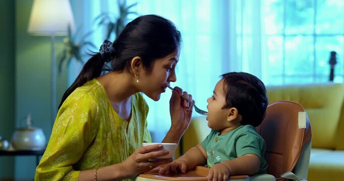 Indian infant eating baby food, mother feeding with spoon while sitting in modern living room, showing love, care, healthy nutrition, bonding, and early parenting moments focused on child&rsquo;s growth
