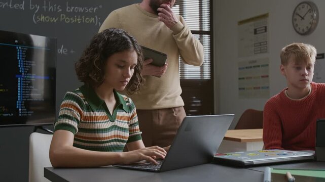 Medium shot of cheerful young Caucasian male teacher walking around classroom, observing schoolchildren writing computer code on laptops, giving advice and correcting mistakes