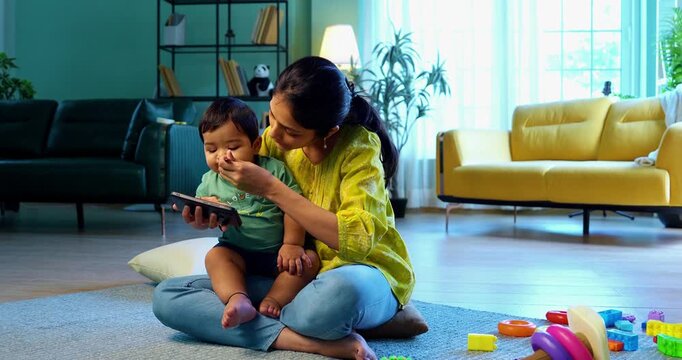 Indian infant eating baby food while watching video on smartphone as mother feeds with spoon, showing modern parenting habits, screen exposure, early tech use, and changing child-rearing practices