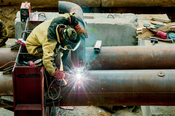 Man welder in full protective gear is welding a large metal pipe on a construction site