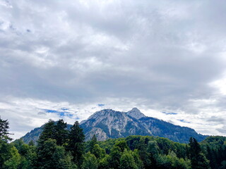 Fototapeta premium An unsurpassed view of alpine meadows at the foot of the forest against the background of sharp peaks of mountain rocks under a cloudy sky on the horizon.