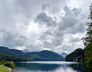 Amazing panorama of the calm emerald surface of the Alpsee mountain lake reflecting the clouds hanging over it surrounded by green forest hills.