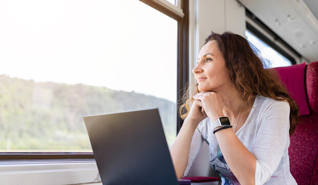 Peaceful middle-aged woman gazing out the train window with a soft smile, resting from work beside her laptop. Reflective moment of relaxation and travel inspiration - Powered by Adobe