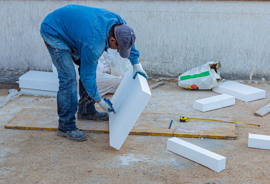 Construction worker cutting a piece of white polystyrene insulation panel (EPS) with a utility knife on the ground, preparing for the external thermal insulation (ETICS) installation process.