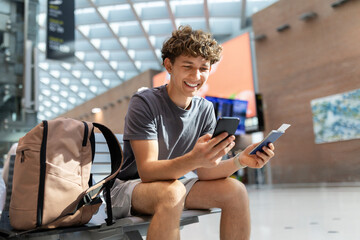 Smiling traveler guy sitting at airport bench using smartphone and holding passport. Happy young man waiting for boarding or booking hotel abroad