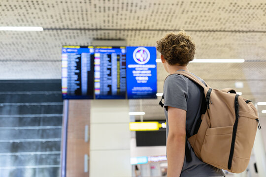 Back view of young traveler with backpack looking at flight board inside airport hall. Journey planning and international departure scene, selective focus