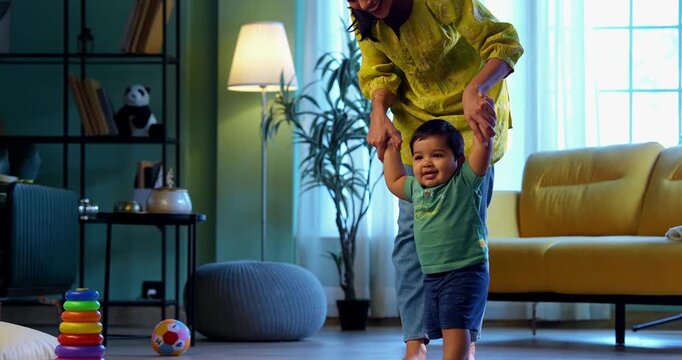 Indian infant boy walking holding young mother's hand in modern living room, learning to balance and take first steps with love, care, affection, and gentle guidance from mom