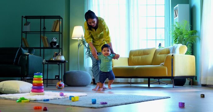 Indian infant boy walking holding young mother's hand in modern living room, learning to balance and take first steps with love, care, affection, and gentle guidance from mom
