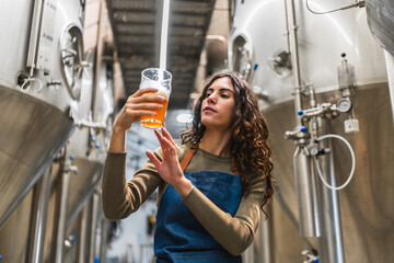 Female brewmaster holding a glass of craft beer, visually examining its quality and color in a modern microbrewery