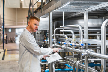 Red haired man in a lab coat checking control valves and industrial piping in a modern production facility, ensuring quality control