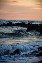 Dynamic ocean waves crashing on dark rocks during a pastel sunset.