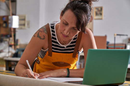 Female artisan planning workshop projects, writing notes, and checking details on a laptop, managing her small business