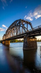 Fototapeta premium A tall, metallic arched structure crosses a wide body of water. Blue skies, with subtle clouds, provide the backdrop. The sunlight casts shadows