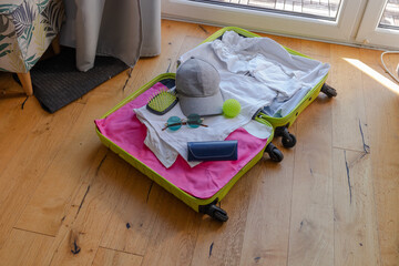 A bright green suitcase is open on a wooden floor. It contains white clothing, a gray hat, sunglasses, a tennis ball, and a blue wallet.