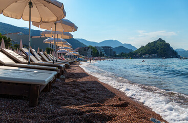 city beach with sun loungers and umbrellas with beautiful coastline and view of Adriatic sea in Petrovac, Montenegro, with high forested hills in the background