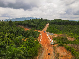 An aerial view of a muddy road under construction in the remote interior of East Kutai, Borneo, Indonesia. It cuts through rainforests and palm oil plantations with karst mountains on the horizon.