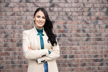 A confident woman smiles against a brick wall, showcasing her professional attire and vibrant personality in an urban environment. Portrait