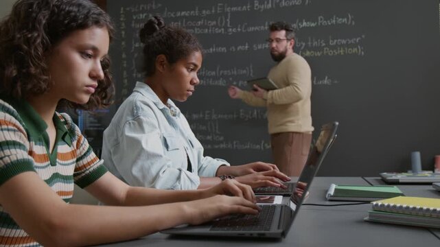 Medium side shot of teenage Caucasian and African American girls sitting at table, writing computer code on laptops during lesson, while male teacher is explaining algorithm on chalk blackboard