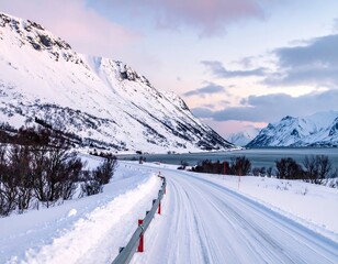 A snow-covered road winds toward a serene fjord surrounded by majestic, snow-laden mountains under a pastel-colored sky at dusk