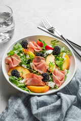 Fresh salad with peaches, arugula, feta, prosciutto and berries in a plate on a light background