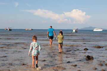 A joyful family scene on the tropical beach of Phi Phi Island, Thailand. A father walks through shallow water with his son and daughter, holding their snorkeling gear as traditional fishing boats floa