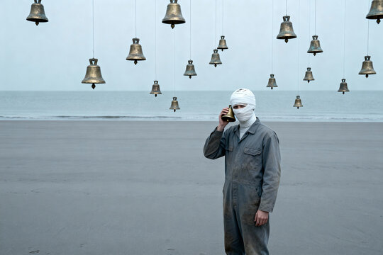 A mysterious figure in overalls with a bandaged head uses a bell as a phone on a deserted beach under a surreal sky of floating bells.