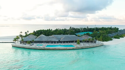 Obraz premium Aerial view of a main resort building with a large pool on an island point, surrounded by white sand and turquoise water at sunset