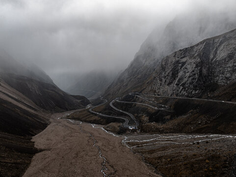 A winding mountain road meanders through a valley with flowing water and heavy mist, set against rugged peaks and dark tones, creating a deep and tranquil natural scene that emphasizes the grandeur