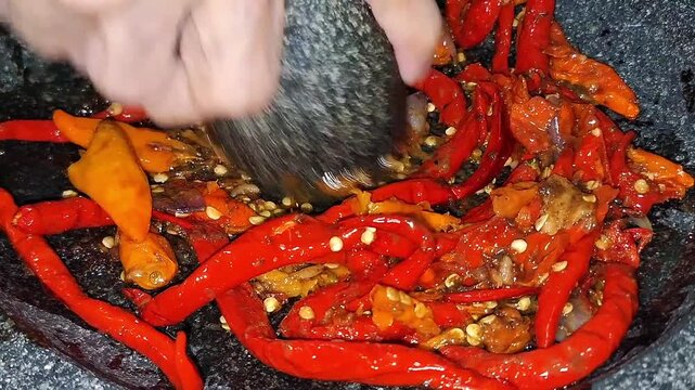 Close-up of red chili, cayenne pepper, and shallots ground on a stone pestle during a traditional Sambal Dadak preparation.