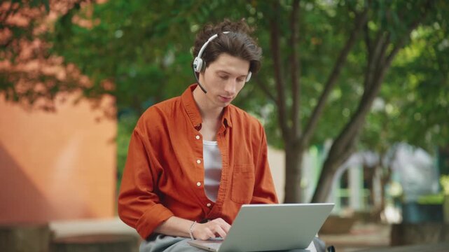 Man wearing headset sitting outdoors using laptop. Focused on online communication while working remotely in customer support role. Calm park setting with green trees in background.