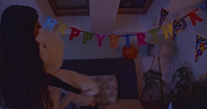 Joyful woman celebrating her birthday at home, playfully dancing and hugging a giant teddy bear. The room is decorated with balloons and happy birthday banners, creating a cozy party atmosphere.