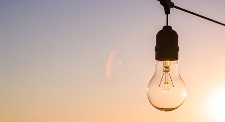 A single unlit light bulb hangs against a beautiful, warm sunset sky. A concept of waiting for an idea, potential, or the end of the day.