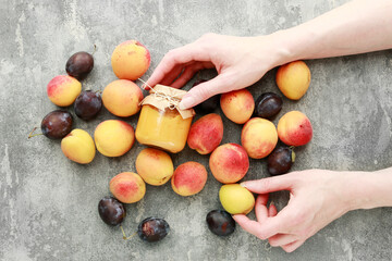 A woman holds a small jar of jam in her beautiful hands against the background of fruit lying on a...