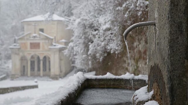 Public drinking fountain along the sacred path of the Sacred Mount of Varese (Santa Maria del Monte) Italy, with an ancient chapel under snow in the background, Unesco Site. Tenth chapel in winter