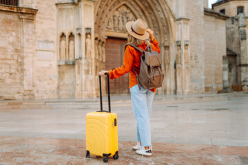 A rear view of female traveler with yellow suitcase and backpack walking through sunlit square....
