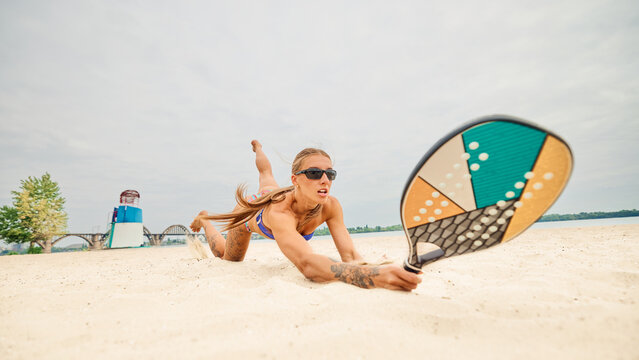 Female padel athlete diving on sand with paddle in stretched hand. Concept of fitness, women health, emotions, social media lifestyle and generational culture.