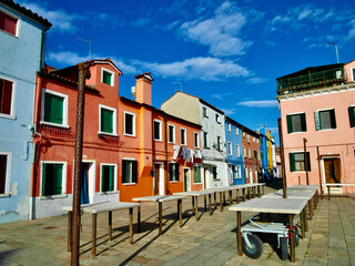 Vibrant colorful houses along a quiet street on Burano Island, Venice, Italy. The pastel facades, laundry lines, and blue sky reflect the charming Mediterranean lifestyle.