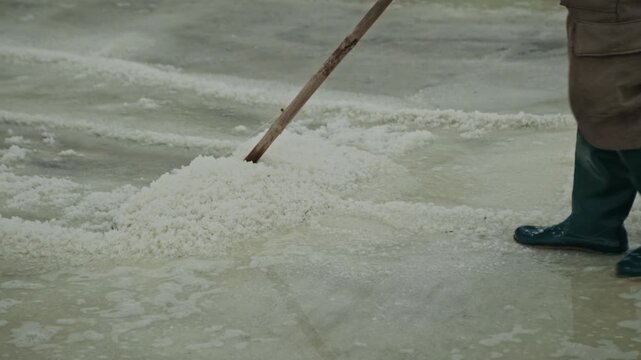 Worker in rubber boots rakes wet salt in pile with wooden tool in evaporation pond closeup. Laborer harvests mineral deposit condiment with old method at countryside. Manual job