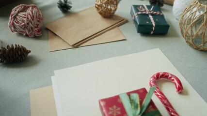 Close up on Christmas gift box wrapped in green ribbon lying next to red and white striped candy cane on tabletop cluttered with envelopes and craft materials, copy space - Powered by Adobe