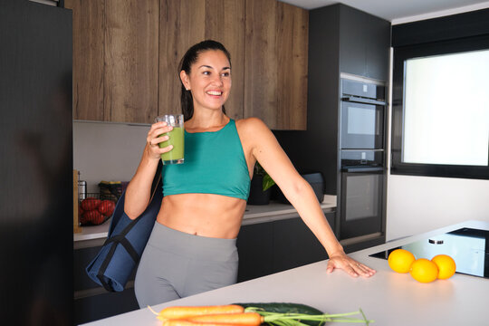 Smiling woman with yoga mat drinking green smoothie in modern kitchen