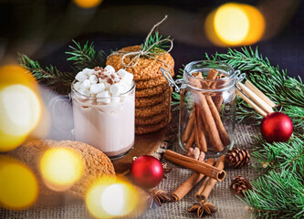 A Christmas photo with marshmallows in a mug and cinnamon sticks, as well as Christmas cookies and fir branches, as well as star anise fir cones and red Christmas tree decorations nearby