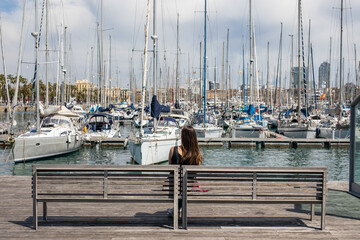 Serene getaway to Barcelona's harbor as a woman enjoys the waterfront view from a bench, yachts bobbing gently in the marina on a beautiful day