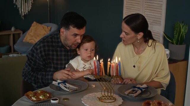 Medium shot of young Jewish parents sitting at heartwarming festive table at home and teaching Hanukkah traditions to their toddler son spinning dreidels and looking at candles burning on hanukkiah