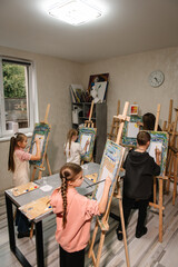 A teacher explains to the children how to draw picture in art class. A group of school children with brushes and an easel in art lesson began drawing landscapes. Children enjoy painting in a workshop.