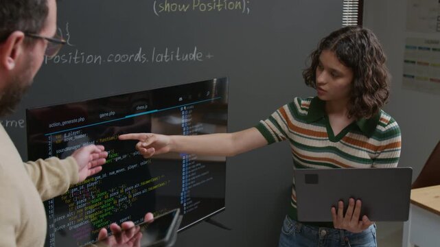 Medium shot of male programming teacher reviewing computer code written by teenage girl on large screen in classroom, pointing out mistakes, answering questions and explaining variables