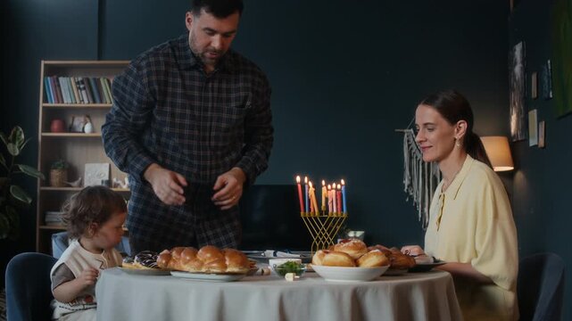 Young Jewish father as head of family reciting blessing holding two loaves of traditional holiday bread challah, then putting it on festive table having heartwarming Hanukkah dinner with wife and son